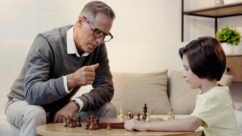παππούς με γυαλιά και έξυπνο εγγόνι που παίζει σκάκι στο σαλόνι - Φωτογραφία, εικόνα