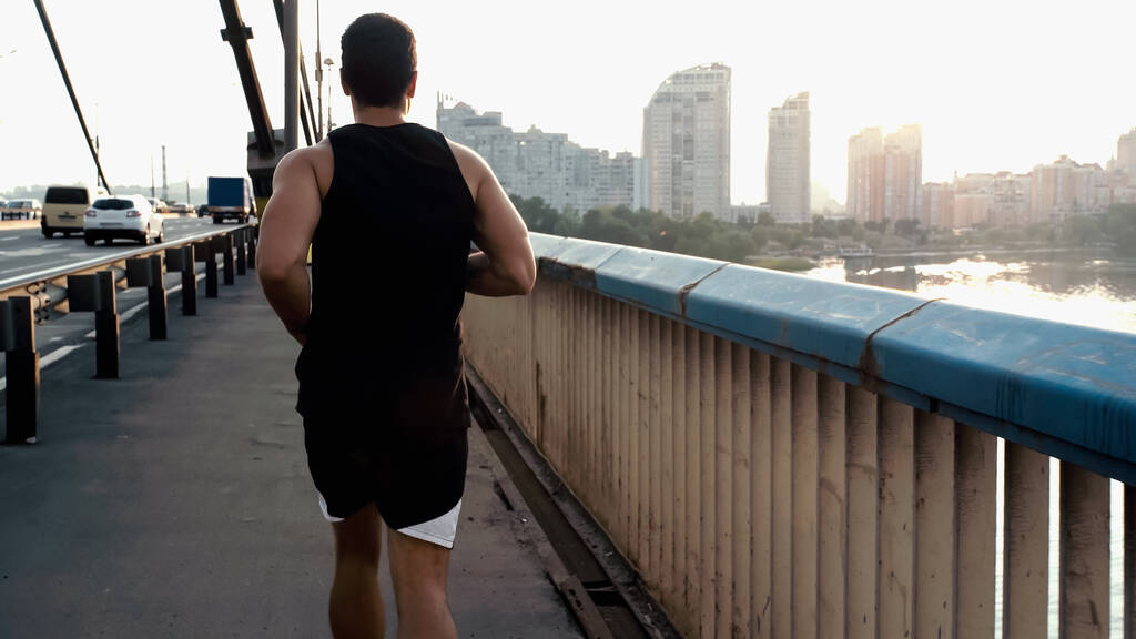 назад вид спортсмена смешанной расы, бегущего по городскому мосту через реку - Фото, изображение