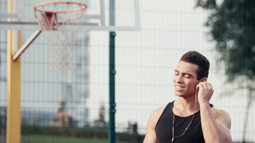joven deportista bi-racial con los ojos cerrados ajustando auriculares al aire libre - Foto, Imagen