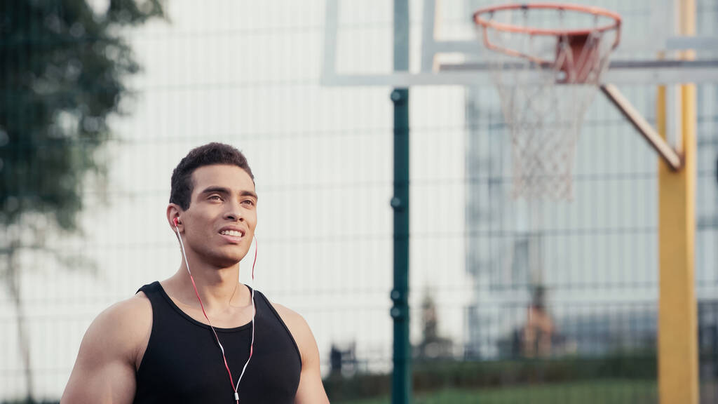 joven deportista de carrera mixta en auriculares mirando hacia otro lado cerca borrosa anillo de baloncesto - Foto, imagen