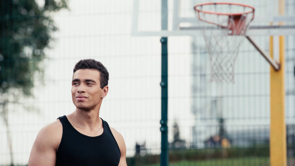 atlético bi-racial homem olhando para longe perto borrado basquete anel ao ar livre - Foto, Imagem