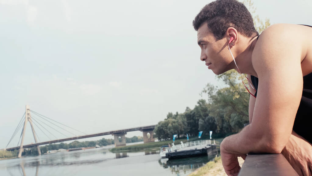 вид сбоку спортивного бирасового человека, слушающего музыку, глядя на реку - Фото, изображение