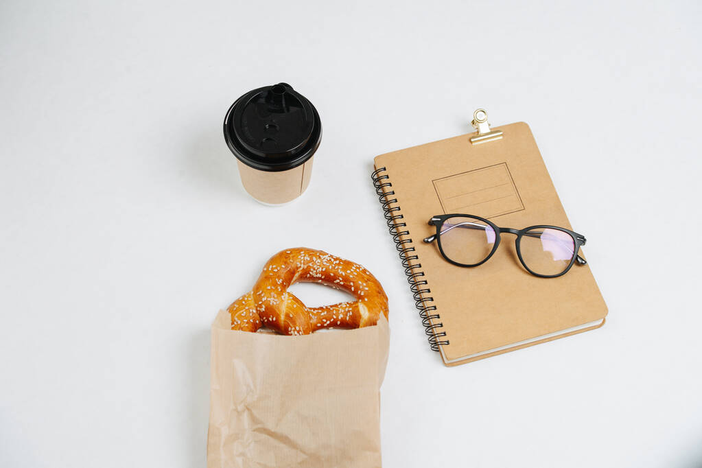 Σύνθεση υψηλής γωνίας ενός φλιτζανιού καφέ, ζαχαροπλαστικής και γυαλιών σε ένα σημειωματάριο. Πάνω από λευκό φόντο. - Φωτογραφία, εικόνα