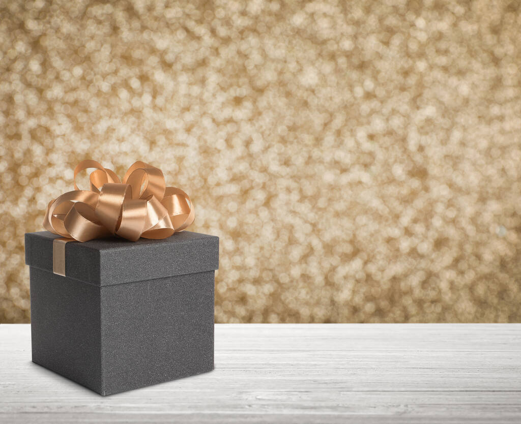 Подарочная коробка на белом деревянном столе на блестящем золотистом фоне, эффект боке. Пространство для текста - Фото, изображение