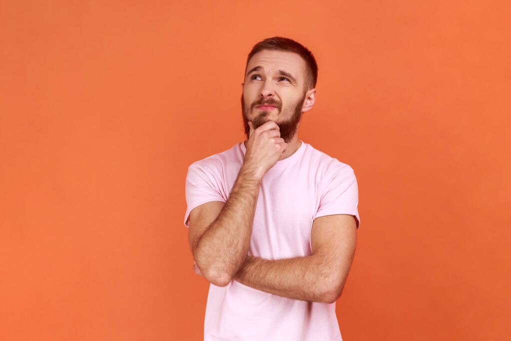 Portrait of thoughtful handsome bearded man holding his chin and pondering idea, confused not sure about solution, wearing pink T-shirt. Indoor studio shot isolated on orange background. - Fotó, kép