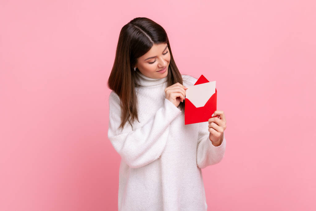 Portrait of happy woman holding red romantic envelope and looking at letter with smile and interest, wearing white casual style sweater. Indoor studio shot isolated on pink background. - Foto, immagini