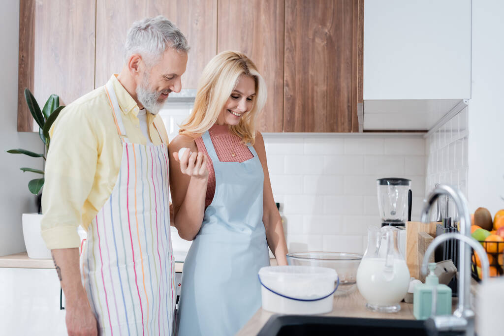 Θετική γυναίκα κρατώντας αυγά κοντά στον σύζυγο και τα συστατικά στην κουζίνα  - Φωτογραφία, εικόνα