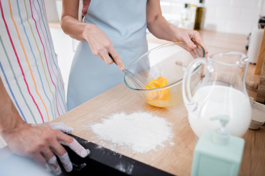 Vista ritagliata di donna che mescola uova in ciotola vicino agli ingredienti e marito in cucina  - Foto, immagini