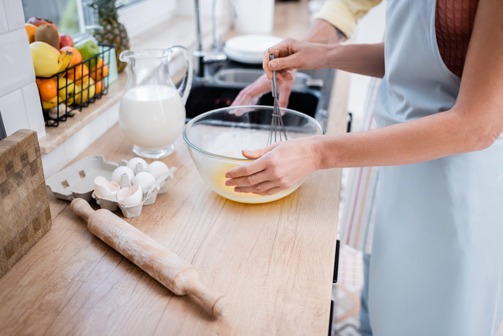 Vista recortada de la mujer en delantal mezclando huevos en un tazón cerca del rodillo y el marido en casa  - Foto, Imagen