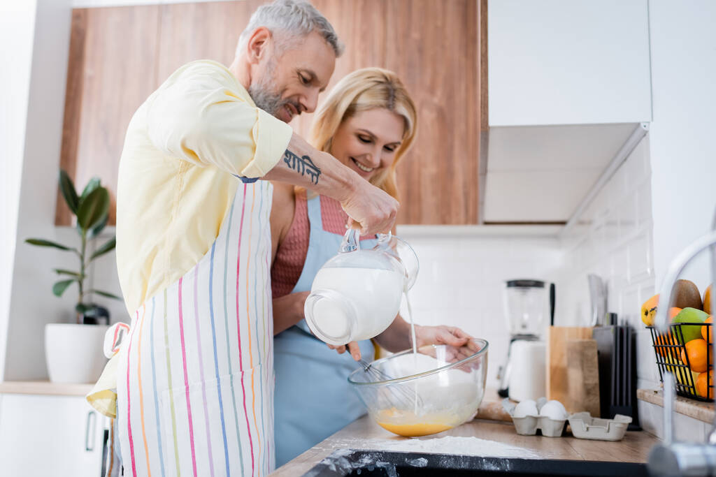Sonriente hombre vertiendo leche en un tazón cerca de la esposa cocinando en la cocina  - Foto, Imagen