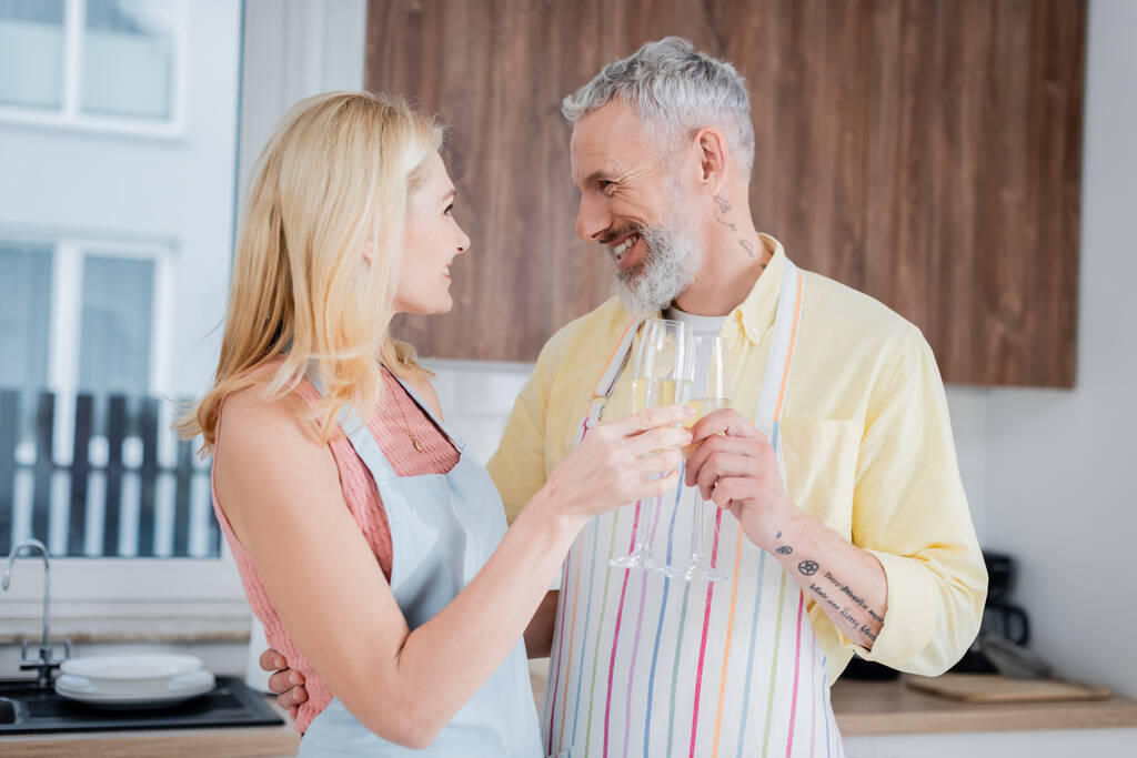 Side view of smiling middle aged man in apron clinking champagne with wife at home  - Photo, Image