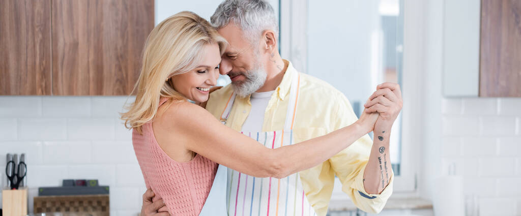 Позитивний зрілий чоловік в фартусі танцює зі усміхненою дружиною на кухні, банер
  - Фото, зображення