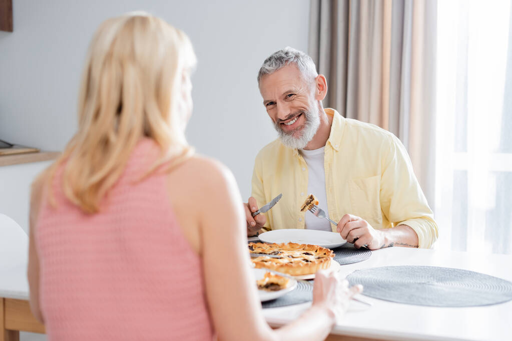 pozytywny Dojrzały mężczyzna patrząc na zamazane żona w pobliżu ciasto w kuchni  - Zdjęcie, obraz