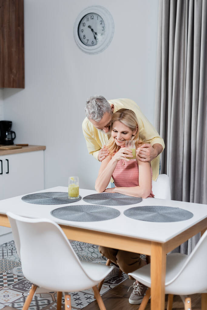 Mature homme étreignant femme souriante avec verre de smoothie dans la cuisine  - Photo, image