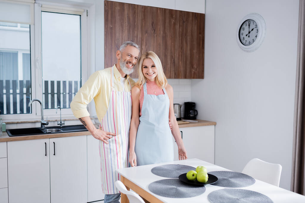 glimlachen volwassen man knuffelen vrouw in schort en kijken naar camera in keuken  - Foto, afbeelding