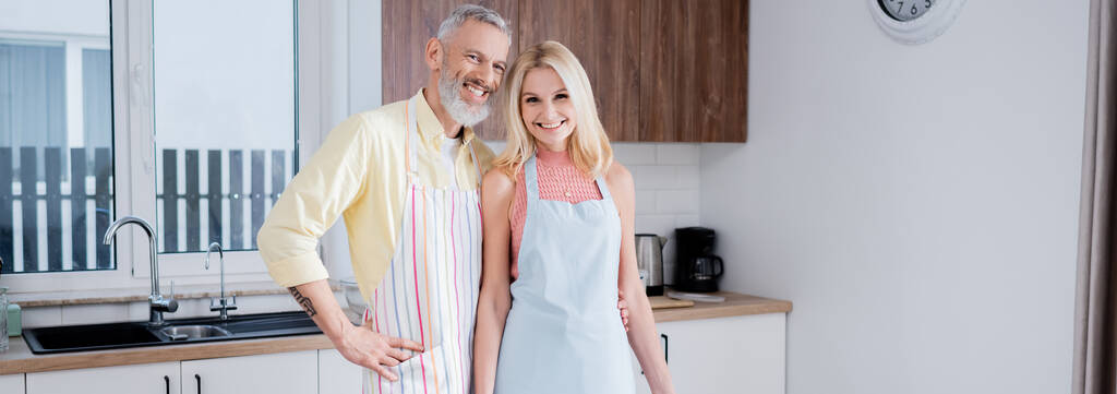 Positive reife Paar in Schürzen Blick auf Kamera in der Küche, Banner  - Foto, Bild
