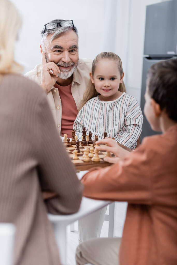 veselý senior muž hraje šachy s manželkou a vnoučaty na rozmazané popředí - Fotografie, Obrázek
