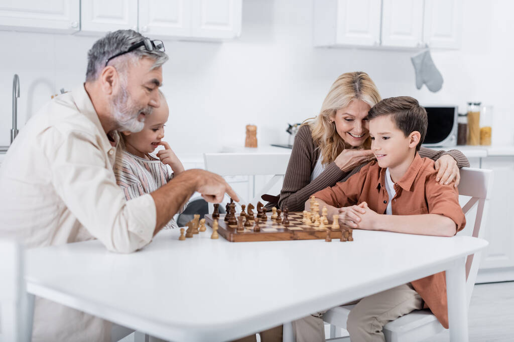 vanhempi mies osoittaa shakkilautaa lähellä lastenlapsia ja vaimo keittiössä - Valokuva, kuva