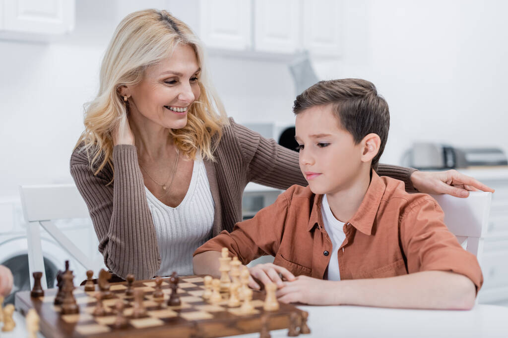 mulher de meia idade sorrindo perto do neto jogando xadrez na cozinha - Foto, Imagem