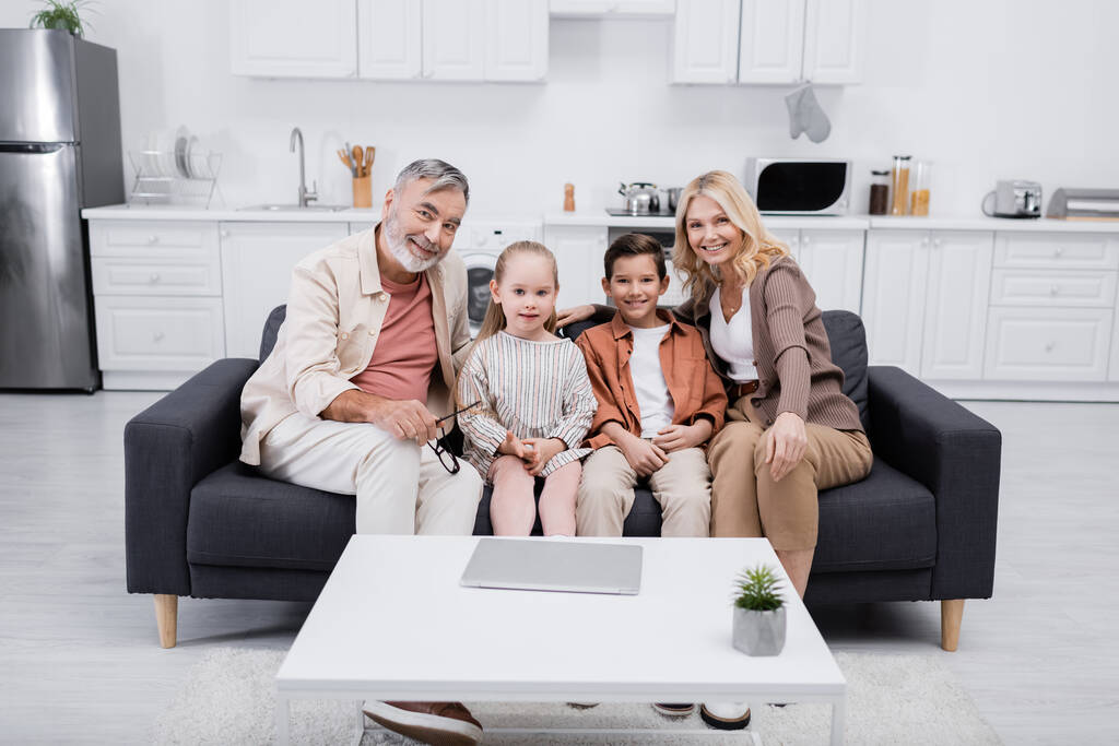 heureux grands-parents avec des enfants regardant la caméra tout en étant assis sur le canapé près d'un ordinateur portable - Photo, image