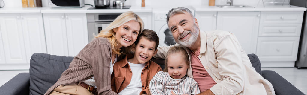 улыбающиеся дети с бабушкой и дедушкой, смотрящие в камеру на диване дома, баннер - Фото, изображение