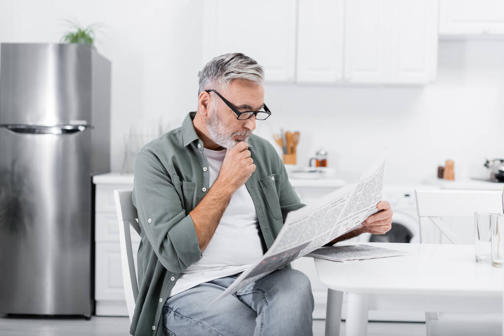 thoughtful senior man in eyeglasses reading morning newspaper in kitchen - Photo, Image