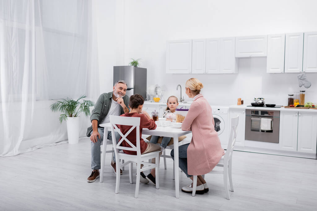 senior man smiling near grandchildren and wife having breakfast in modern kitchen - Photo, Image