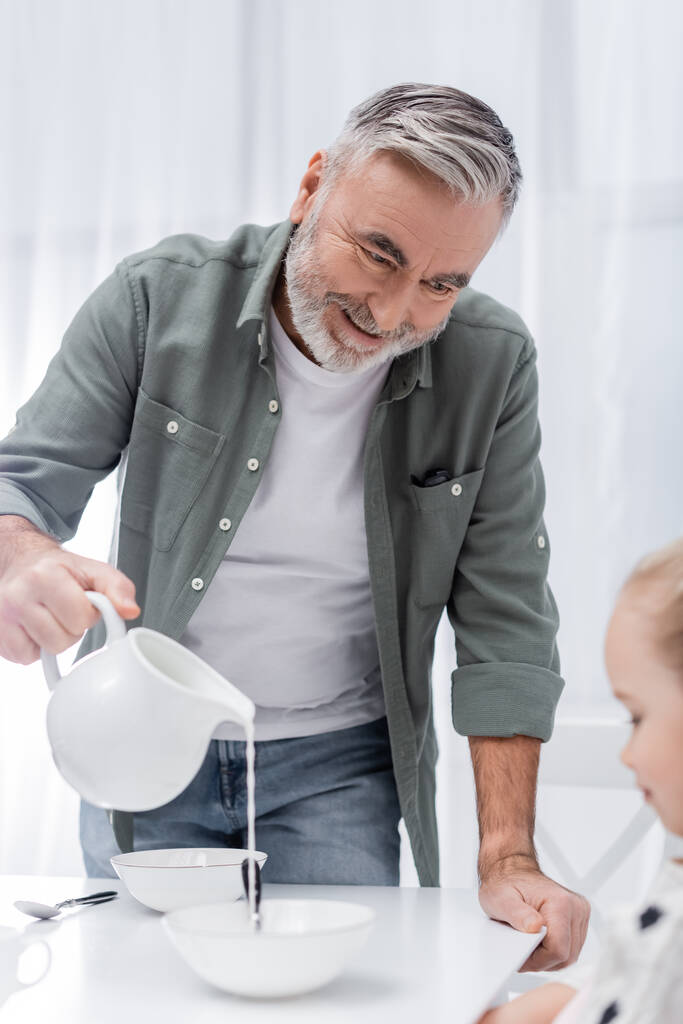 glücklicher Opa schüttet frische Milch neben verschwommenem Mädchen in Küche - Foto, Bild