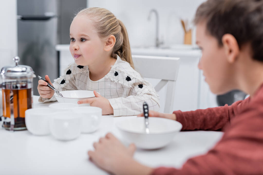 girl having breakfast near blurred brother in kitchen - Photo, Image