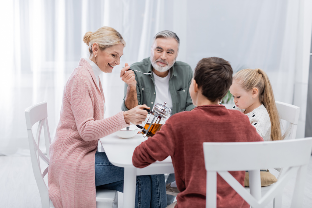 sonriente mujer vertiendo leche cerca feliz marido y nietos durante el desayuno - Foto, imagen