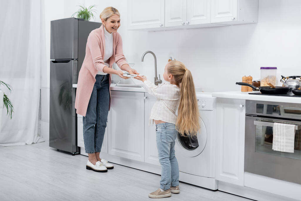 Маленькая девочка дает тарелку улыбающейся бабушке, стоящей возле раковины на кухне - Фото, изображение