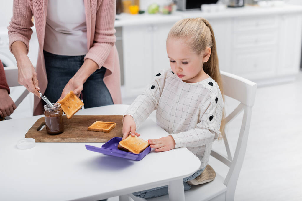 жінка готує тости з шоколадною пастою біля онуки з обідньою коробкою на кухні
 - Фото, зображення