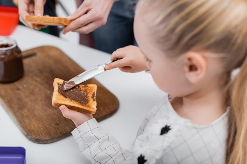 fille floue étaler la pâte de chocolat sur le pain grillé tout en aidant mamie préparer des sandwichs - Photo, image