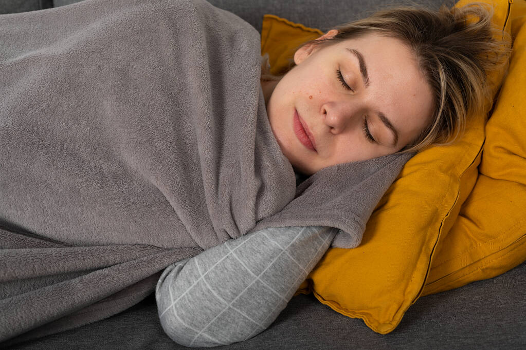 Νεαρή γυναίκα με την εποχιακή γρίπη έχει πονοκέφαλο και πυρετό, ξαπλωμένη στον καναπέ τυλιγμένο σε ένα balnket  - Φωτογραφία, εικόνα