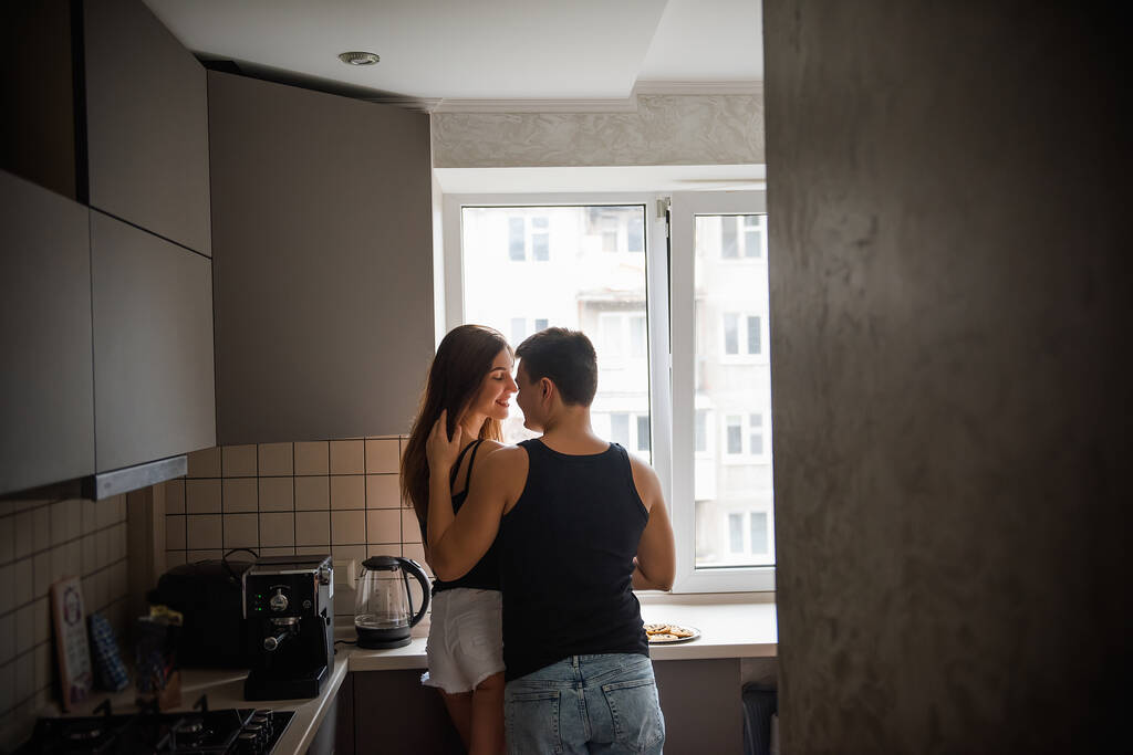 Jeune couple de milléniaux en t-shirts noirs, jeans serrant dans la cuisine le matin par la fenêtre. Amoureux amoureux s'amusent avant le petit déjeuner à la maison. L'homme caresse les cheveux des femmes. Style de vie vie appartement vie - Photo, image