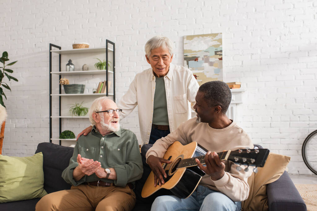 Positieve multi-etnische mannen op zoek naar Afrikaanse Amerikaanse vriend spelen akoestische gitaar thuis  - Foto, afbeelding