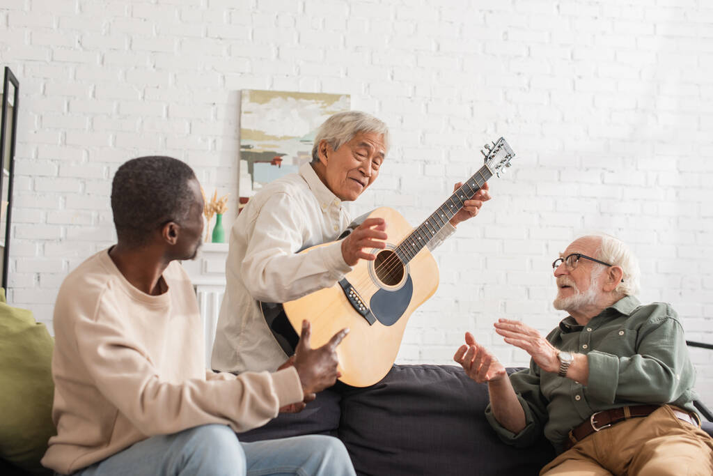 Літні багатоетнічні друзі розмовляють з азіатським чоловіком з акустичною гітарою вдома
  - Фото, зображення