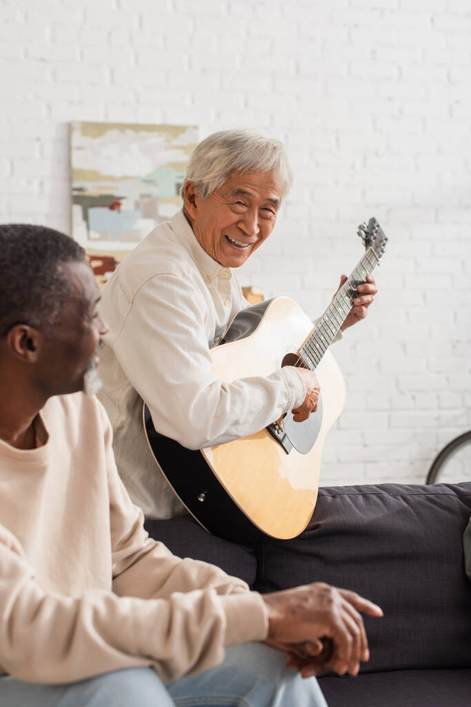 Lächelnder asiatischer Mann spielt Akustikgitarre neben verschwommenem afrikanisch-amerikanischem Freund zu Hause  - Foto, Bild