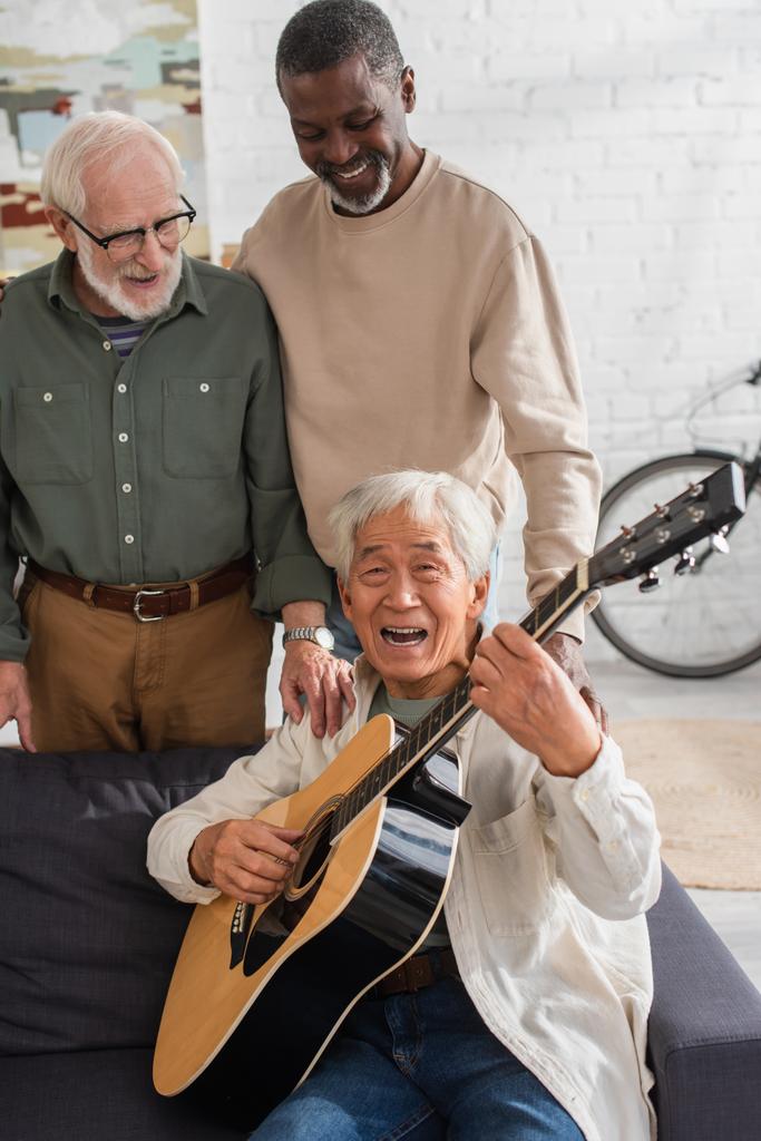Sénior asiático homem tocando guitarra acústica e cantando perto de amigos multiétnicos em casa  - Foto, Imagem