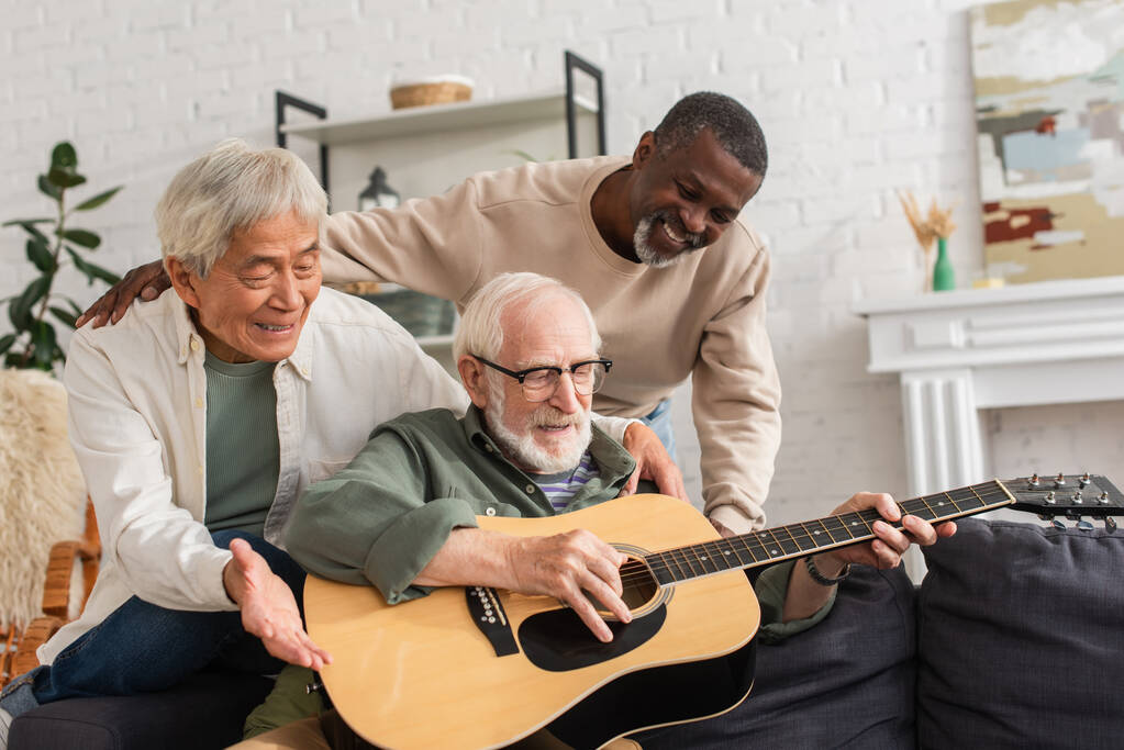 Pozytywni emeryci międzyrasowi patrzą na przyjaciela grającego na gitarze akustycznej w domu  - Zdjęcie, obraz