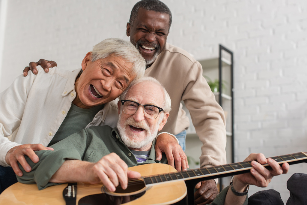 Happy senior interracial friends playing acoustic guitar and looking at camera at home  - Photo, Image