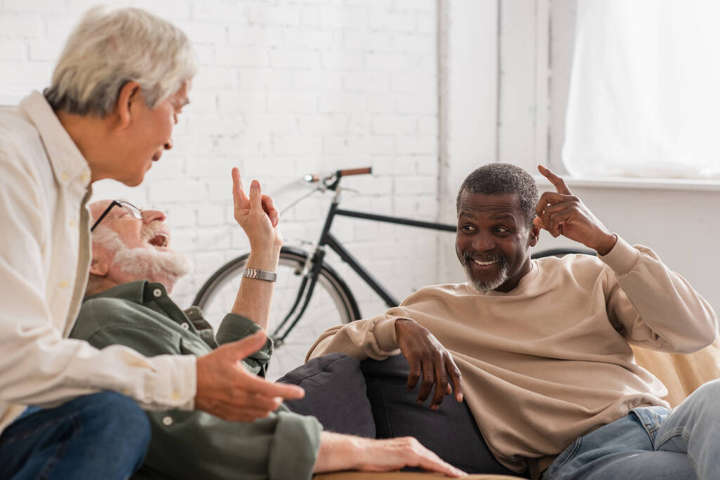 Веселые мультикультурные пожилые мужчины разговаривают дома на диване  - Фото, изображение