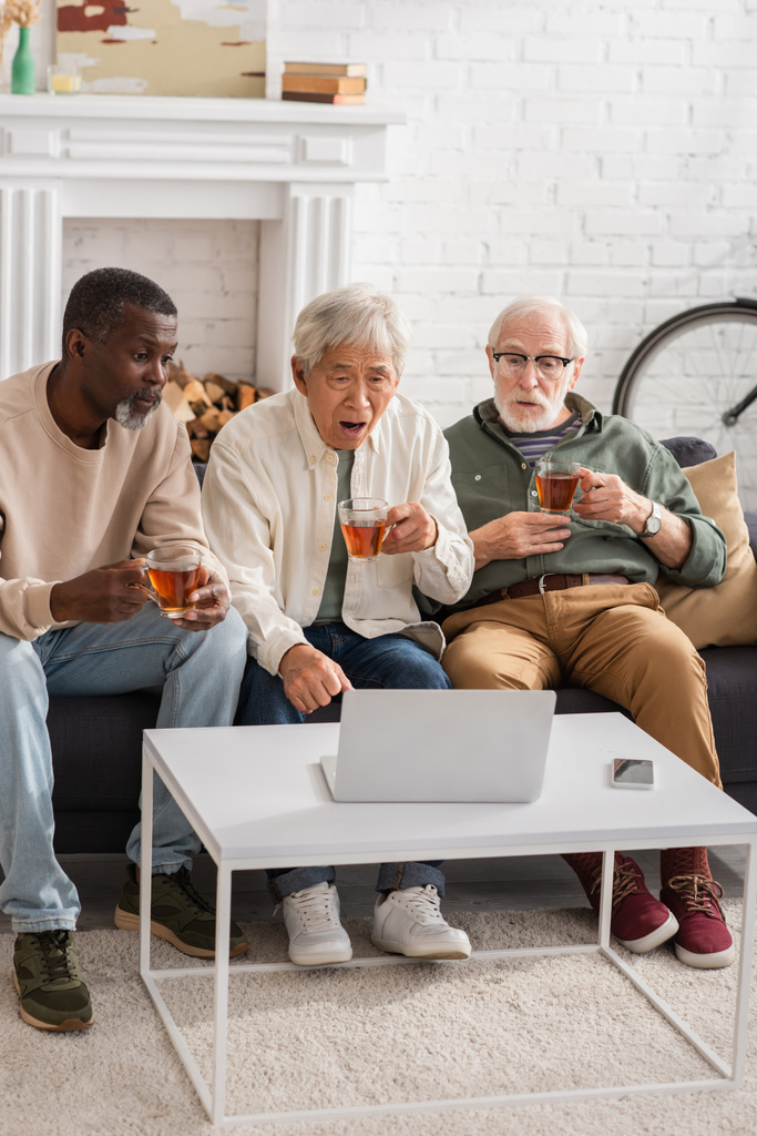 Шокированные мультиэтнические мужчины, держащие чай и глядящие на ноутбук дома  - Фото, изображение