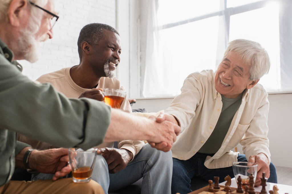 チェスボードの近くで握手をする異人種間の年金受給者と自宅でお茶とアフリカ系アメリカ人の友人  - 写真・画像