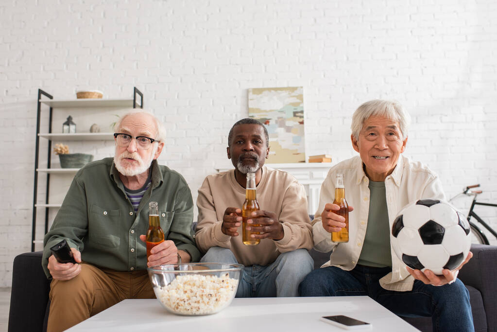 Senior multiethnic friends holding beer and football near popcorn at home  - Photo, Image