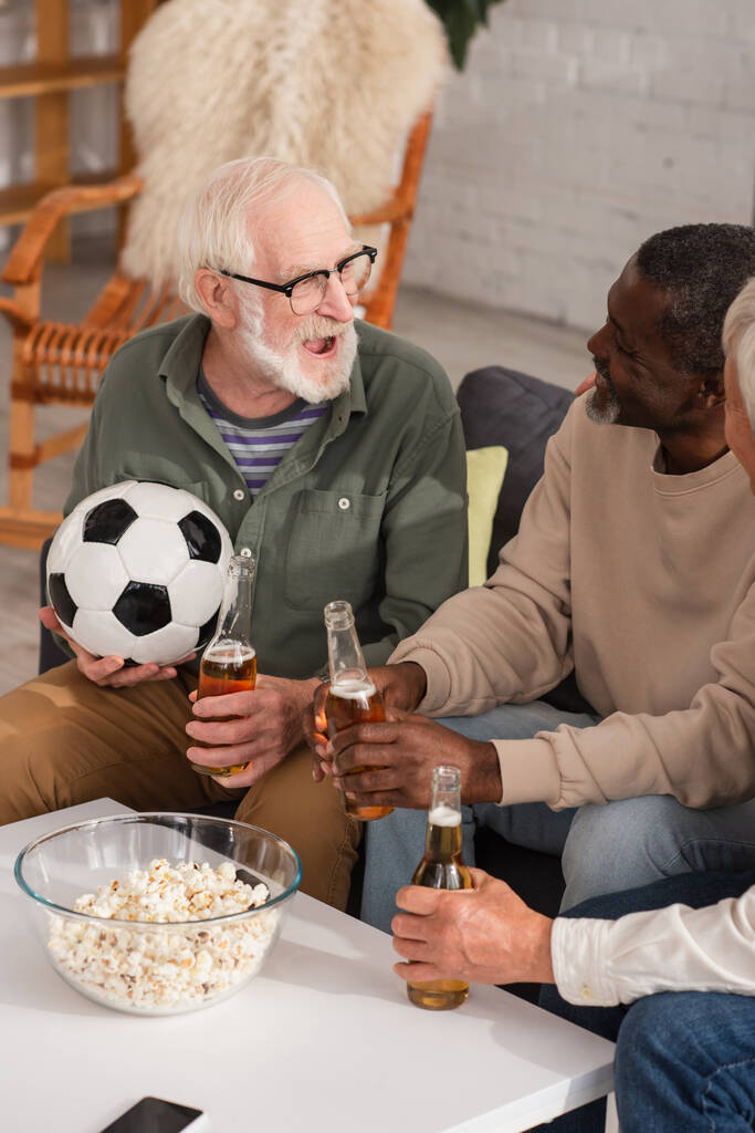 Usmívající se mezirasoví muži s pivem při pohledu na přítele drží fotbal doma  - Fotografie, Obrázek