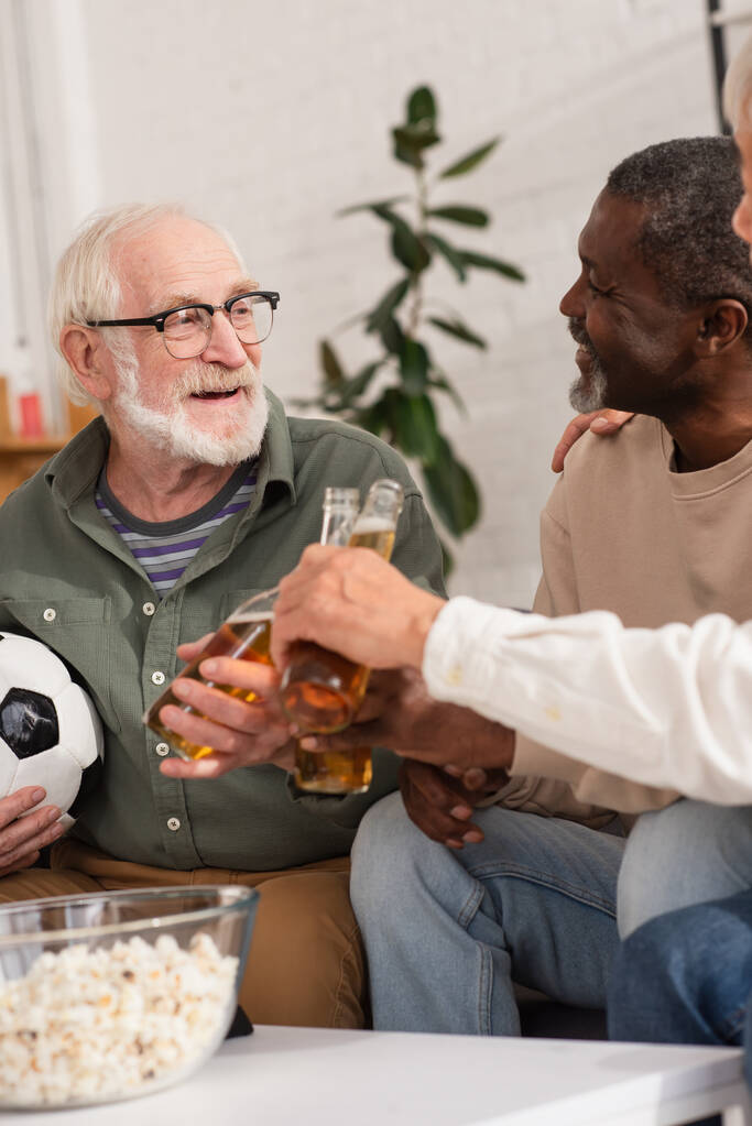Elderly multiethnic men clinking beer with friend holding football at home  - Photo, Image