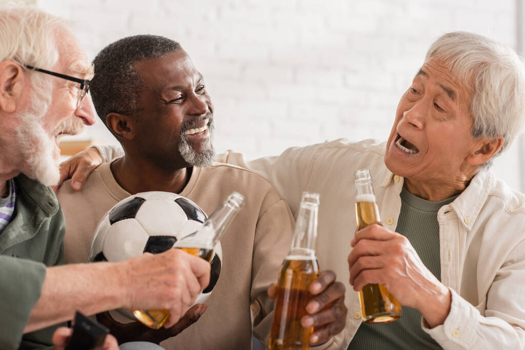 Podekscytowani międzyrasowi starsi przyjaciele trzymający butelki piwa i piłkę nożną w domu  - Zdjęcie, obraz
