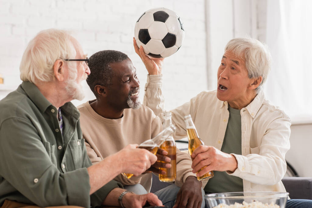 Asian man holding football near multiethnic friends with beer at home  - Photo, Image