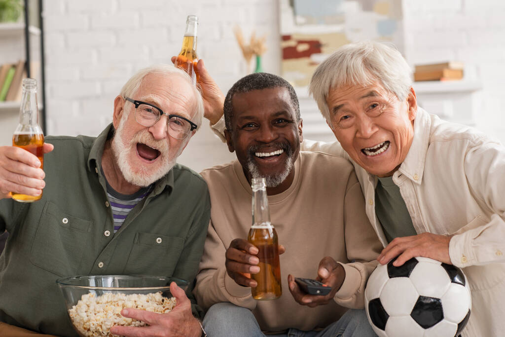 Mosolygó fajok közötti idősebb barátok sör, popcorn és távirányító nézi kamera otthon - Fotó, kép